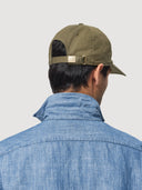 Rear view of menswear model wearing a green Coconut Residence cap and blue chambray shirt on a light grey background
